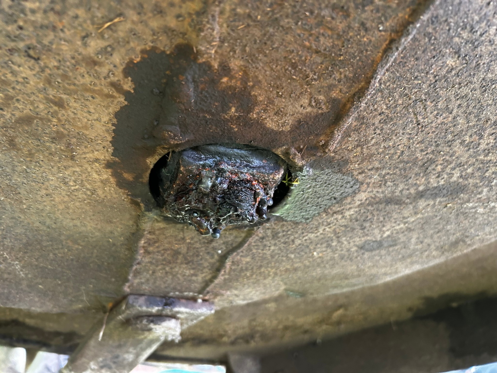 Close-up view of a heavily rusted and corroded metal object, possibly part of a machine or piece of equipment. A dark, viscous substance is dripping from a central hole or fitting. The overall condition suggests significant age, neglect, or exposure to the elements.