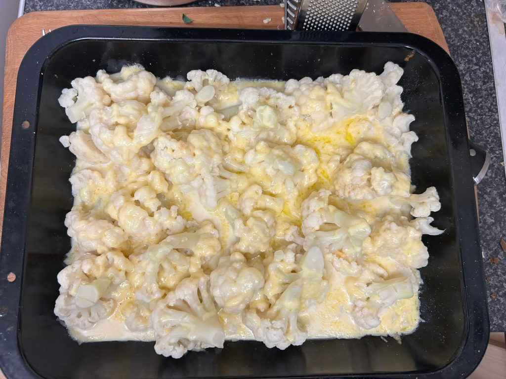 Black baking dish filled with cauliflower florets covered in a creamy, cheesy sauce. The sauce appears to be melted cheese, possibly with a béchamel base. The cauliflower is arranged somewhat loosely in the dish, and some of the cheese sauce has pooled between the florets. The overall appearance suggests a prepared dish, possibly ready for baking or already baked.