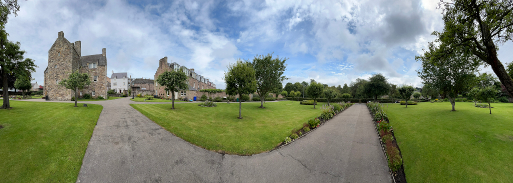 Panoramic view of a well-maintained green lawn, with two curving paved pathways leading towards a stone building, possibly a historic house or castle, nestled amongst other buildings in the background. Mature trees and neatly landscaped flowerbeds are interspersed throughout the lawn, contributing to a tranquil and serene atmosphere. The sky is partly cloudy, suggesting a pleasant day. The scene appears to be in a park-like setting, possibly a historical site or a private estate.