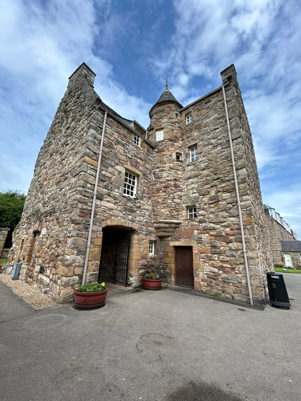 Low-angle view of a historic stone building, possibly a tower house or part of a castle. The structure is made of rough, multi-colored stone, with small windows and a central, conical tower. There's a gated entrance and a dark wooden door visible. Planters with flowers sit near the entrance. Part of a modern building is visible in the background, suggesting the historic structure is in a contemporary setting. The sky is partly cloudy.