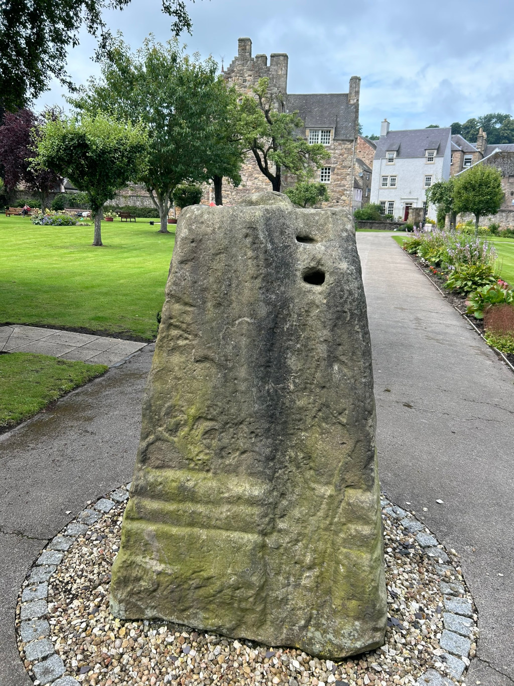 Large, weathered stone standing upright in a grassy area. The stone is irregularly shaped and appears ancient, possibly with carved markings or natural erosion patterns, and it has two noticeable holes near the top.  Behind it is a paved walkway leading toward a stone building that looks like an old manor house or castle, with other buildings visible in the background. The overall setting appears to be a park or garden, with well-maintained lawns and flowerbeds.