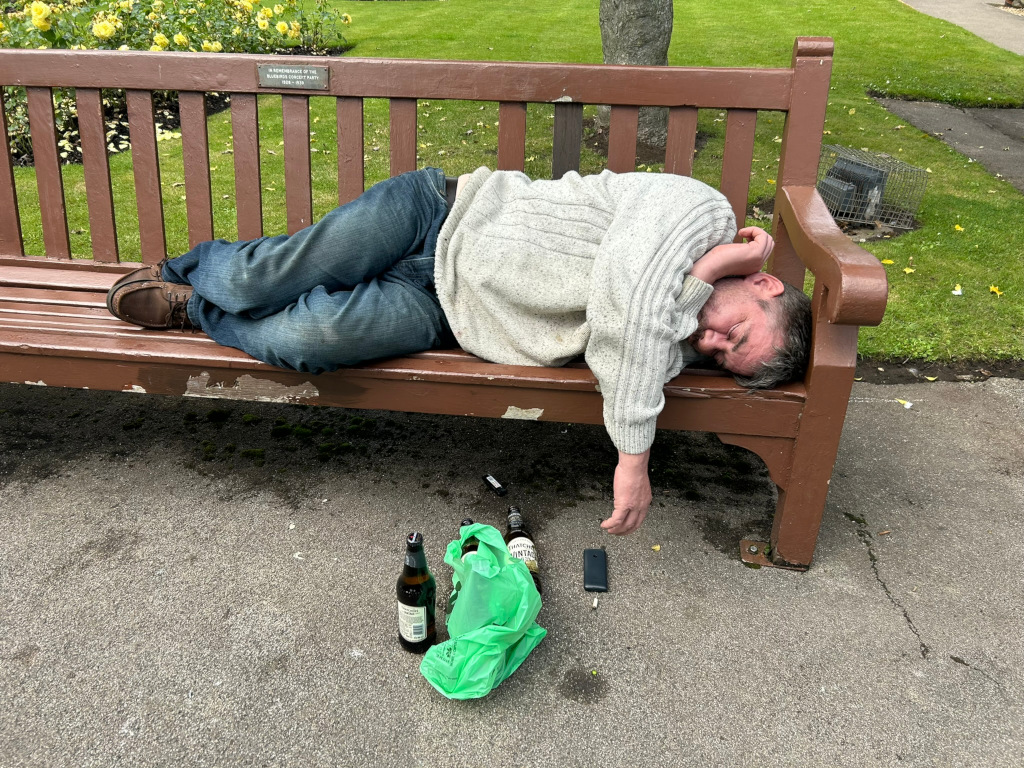 Man asleep on a park bench.  He appears intoxicated, as several empty and partially empty beer bottles are next to him on the ground, along with a discarded plastic bag. His posture suggests he is heavily inebriated or possibly unwell. The scene is set in a park-like setting with greenery visible in the background.