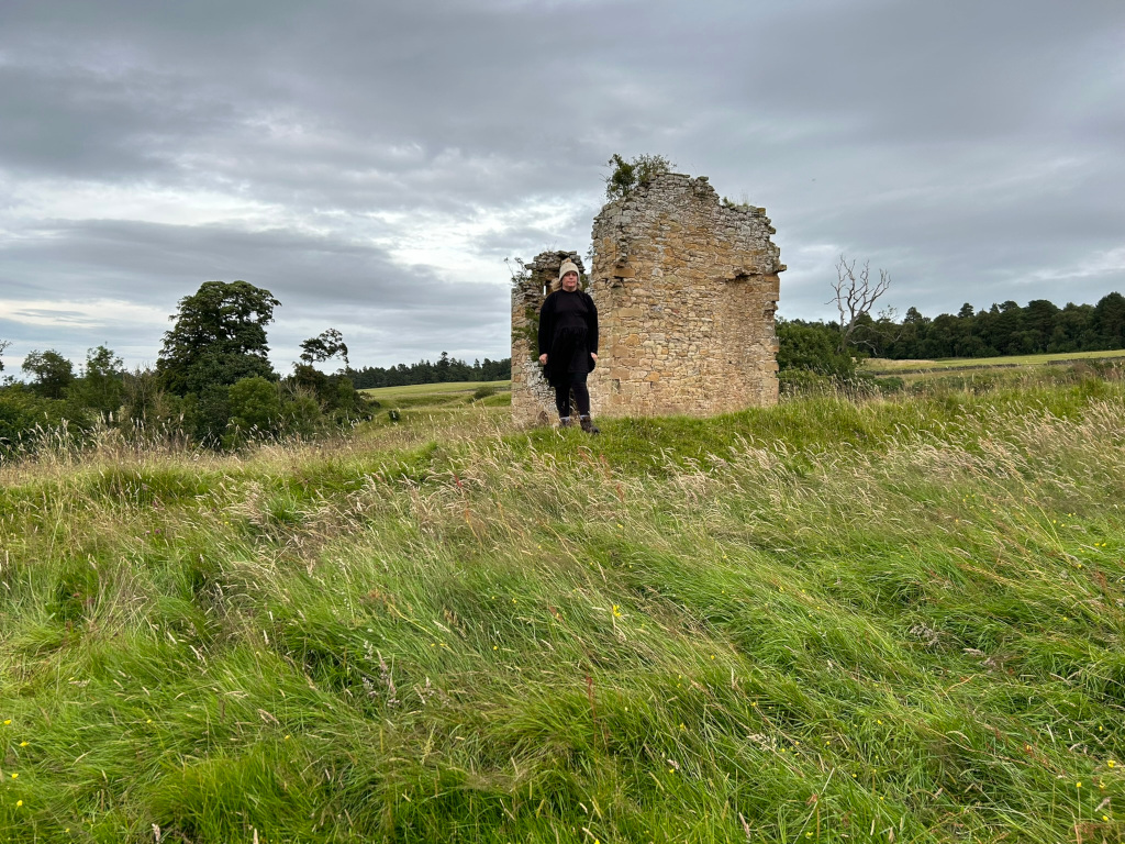 Leonie is standing in front of the ruins of a stone structure, possibly a tower or part of a castle. The ruins are partially crumbled and overgrown with vegetation. She is dressed in dark clothing. The surrounding landscape is a field of tall grass under a cloudy sky. The overall impression is one of age, history, and a sense of solitude or contemplation.
