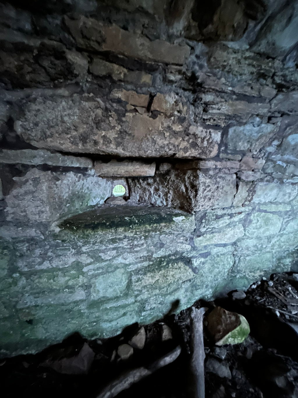 Close-up view of a stone wall, specifically what appears to be the interior of a stone structure, possibly a ruin or an old building. A small, roughly hewn opening or window is visible in the wall, allowing a glimpse of the exterior. The stones are weathered and show signs of age and moss growth, contributing to an overall dark and atmospheric feel. The bottom of the image shows loose stones and debris at the base of the wall.