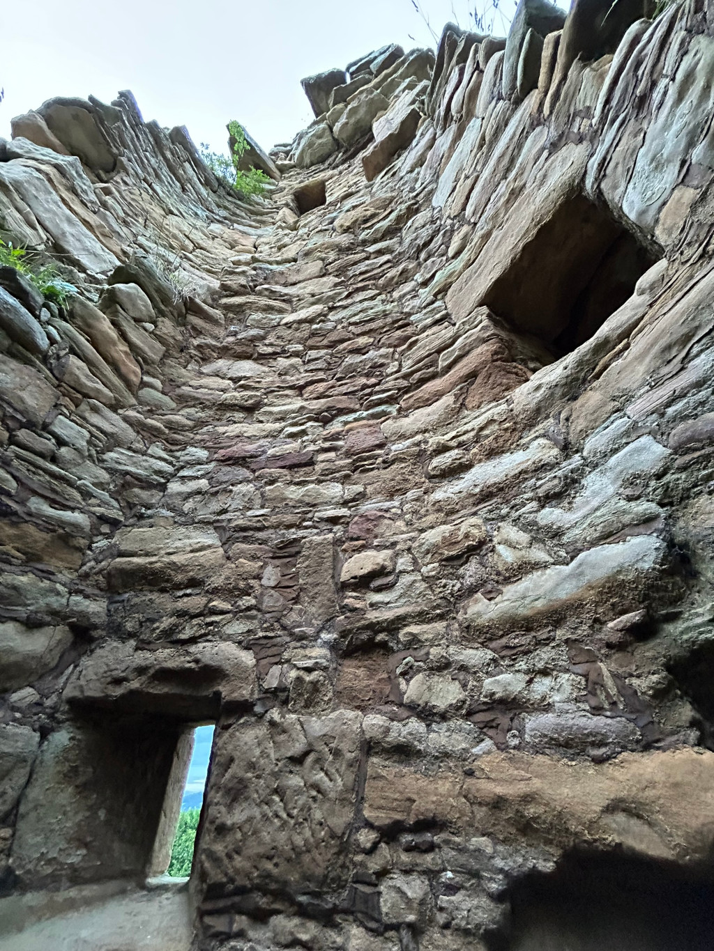 Interior of a circular stone tower, viewed from the bottom looking upwards. The walls are made of rough, irregularly shaped stones of varying shades of brown, grey, and beige. Several small, rectangular openings are visible in the wall, likely former windows or arrow slits. The stones appear ancient and weathered, suggesting a significant age and possibly a state of ruin. A small opening near the bottom allows a glimpse of the outside sky and landscape.