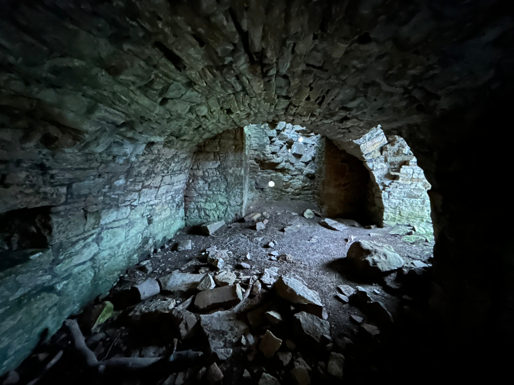 Interior of a crumbling stone structure, possibly a ruin. The space is dimly lit, revealing a low, arched chamber filled with rubble and debris. The stone walls are aged and weathered, showing signs of significant deterioration. A small amount of light enters from an opening further inside the structure, casting shadows and highlighting the texture of the stones.