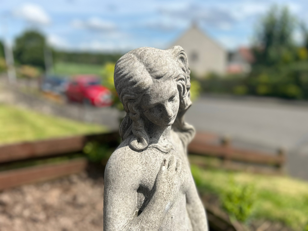 Close-up of a stone statue of a woman, seemingly a garden ornament. The woman is depicted from the chest up, her head slightly bowed, and her hand resting near her chest. The background is blurred, showing a residential setting with houses, greenery, and parked cars, suggesting an outdoor location. The statue appears weathered, showing the texture of the stone.