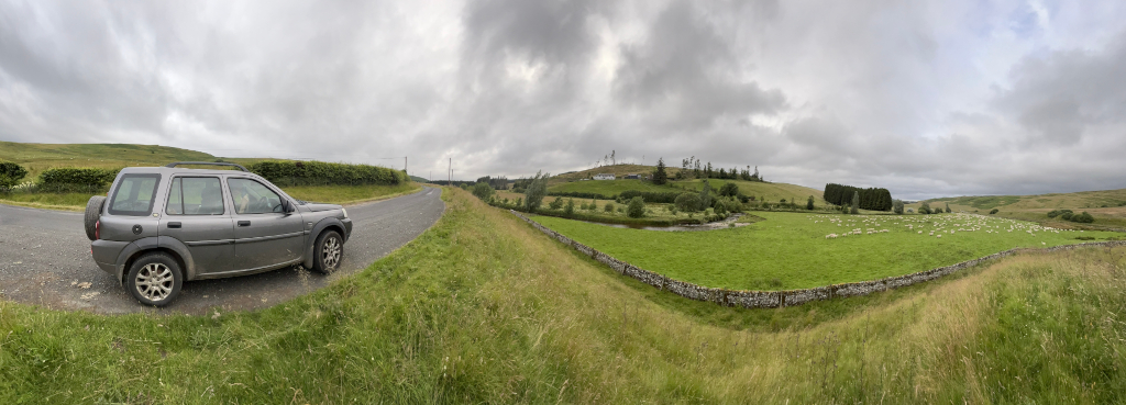 Panoramic view of a rural landscape. A silver Land Rover is parked on the side of a road, which curves gently around a grassy hill. Across the road, a field stretches out, populated by a large flock of sheep. A small stream is visible in the middle distance, beyond which are more fields and a small cluster of buildings, possibly a farm. The sky is overcast.