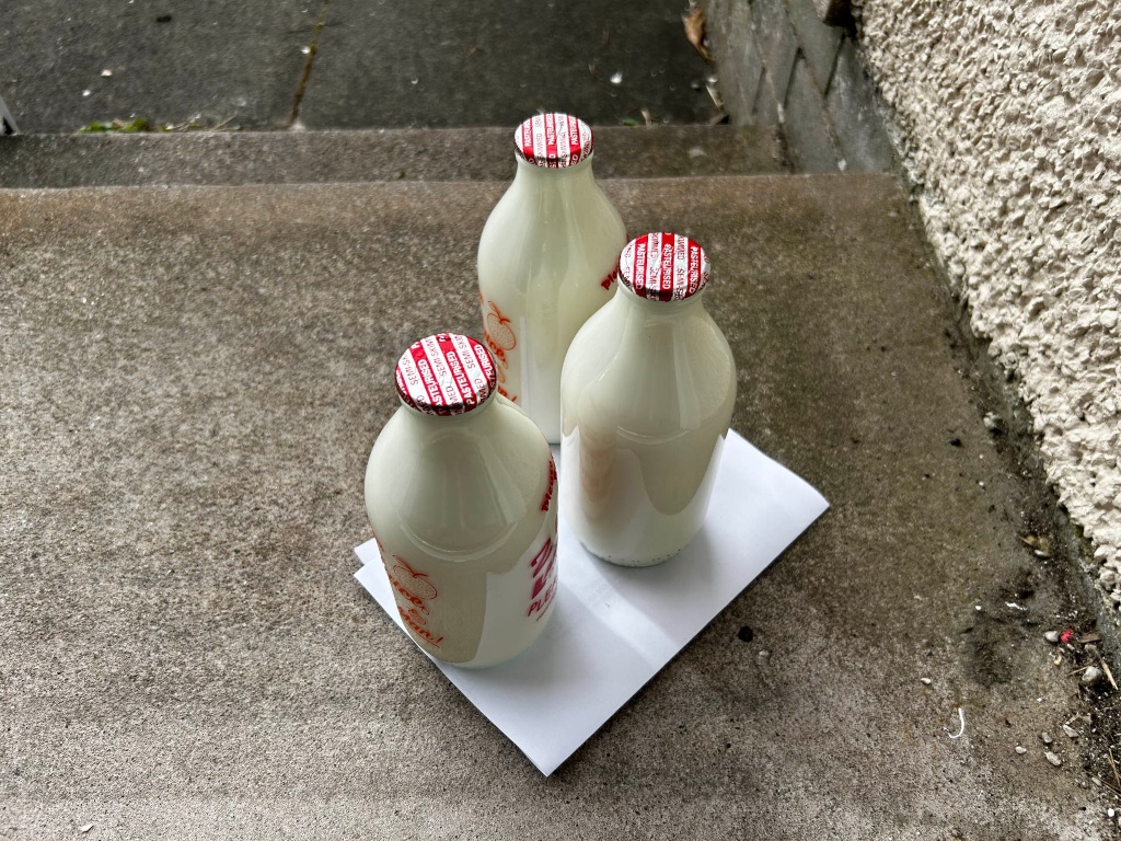 Three glass bottles of milk sitting on a piece of white paper on a set of concrete steps outside a house. The bottles are similar in size and shape, and they all have red and white tamper-evident seals on their caps. The overall impression is of a simple, everyday delivery or arrangement.