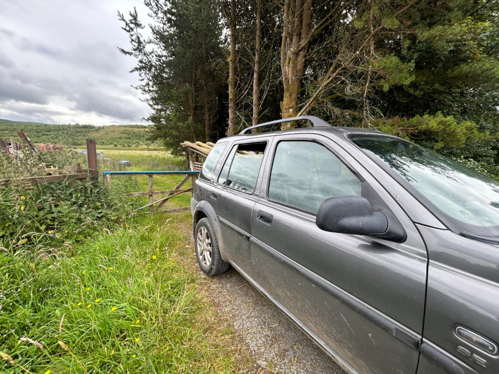 Dark grey Land Rover Discovery parked next to a rustic wooden gate. The car is loaded with several long, light-coloured poles or pieces of lumber on its roof rack. The setting appears to be a rural or countryside area with overgrown vegetation, trees, and a field visible in the background. The overall mood is one of quiet rural life, possibly suggesting a task involving transporting materials in a country setting.