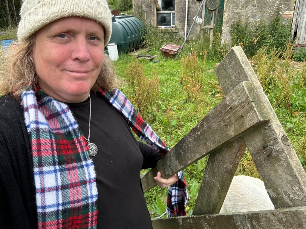 Leonie with shoulder-length blonde hair, wearing a beige knit hat and a black shirt. She's also wearing a red, white, and green plaid scarf and a silver necklace with a circular pendant. She is holding onto a weathered, rustic wooden gate or fence in a somewhat overgrown yard. Part of a white animal, possibly a sheep or goat, is visible through the gate. In the background, there is a stone building and a dark-coloured water tank.