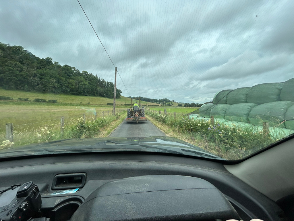 Inside a car driving down a country road.  In the distance, a tractor with a small trailer is visible, moving in the same direction. On the right side of the road, a large stack of wrapped hay bales is prominent. The overall setting is rural, with green fields and trees in the background. The sky is overcast and somewhat gloomy.