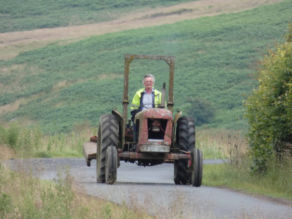 Charlie driving a vintage, rusty red tractor down a country road. The road is flanked by green hills and vegetation. The overall impression is one of rural life and perhaps a sense of peaceful solitude or nostalgia.