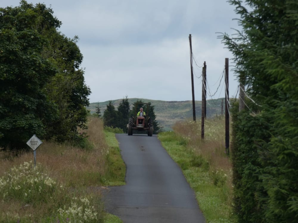 Charlie driving a vintage tractor up a narrow country road.  The road is lined with tall grass and trees, and in the distance, there are rolling hills. A small sign indicating a passing place is visible on the left side of the road.