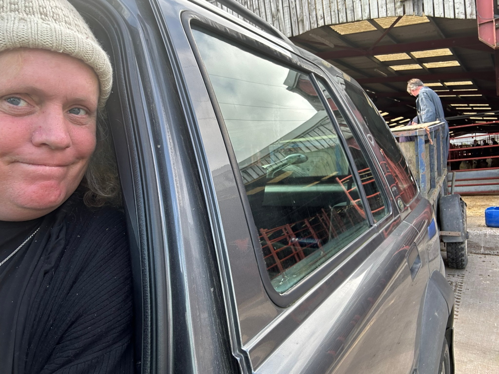 Leonie's face and upper body close-up from inside a dark-grey SUV. She is wearing a cream-coloured knit hat. The background shows Charlie working in a barn or livestock facility, unloading what appears to be livestock from a trailer attached to the vehicle. The Leonie's expression is neutral to slightly sombre.