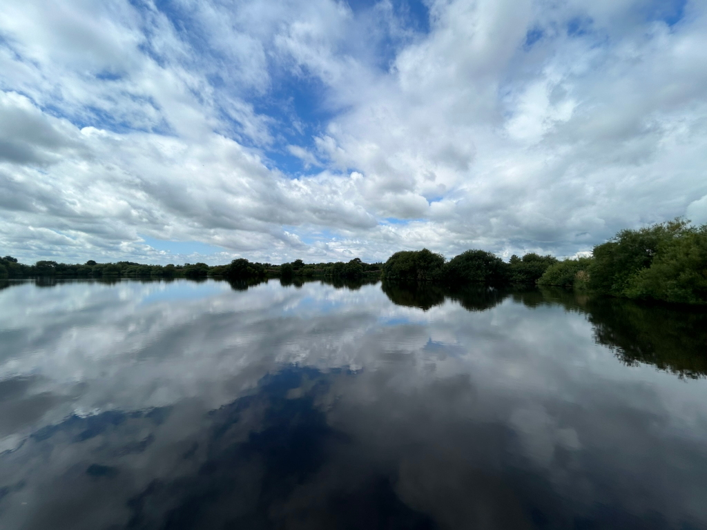 Serene lake scene. The calm water perfectly reflects a partly cloudy sky, creating a nearly symmetrical composition. A line of dark green trees borders the far side of the lake. The overall mood is peaceful and contemplative.
