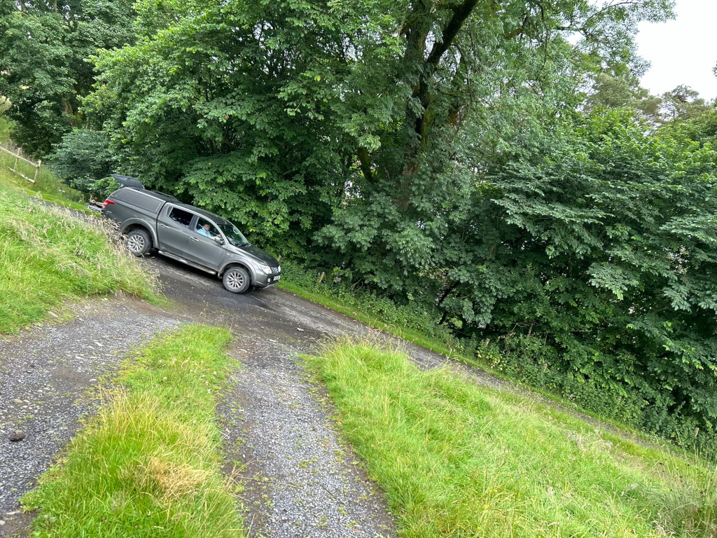 Grey pickup truck parked on a gravel road. The road is bordered by lush green grass and dense foliage. The overall setting appears to be a rural or wooded area. The lighting suggests it's daytime. The truck is parked slightly off-centre, with the perspective drawing the viewer's eye to the vehicle and its surrounding tranquil environment.