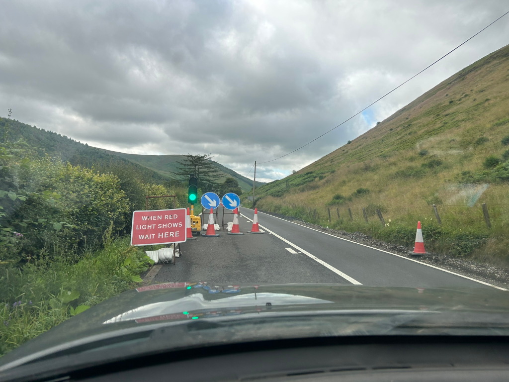Road in a mountainous area, with roadworks in progress. Traffic cones and a temporary traffic signal are visible, along with a sign instructing drivers to wait when the red light is showing. The overall mood is functional, depicting a momentary pause in travel due to construction. The background features green hills under a cloudy sky. The perspective is from inside a vehicle, looking down the road towards the construction site.