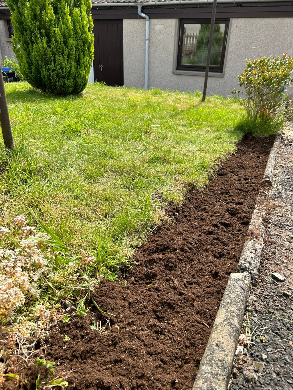 Section of garden where the turf has been removed, revealing dark brown soil. The soil is adjacent to a lawn and runs alongside a curb separating the lawn from a paved area. A small, well-maintained evergreen bush is visible in the background, along with a section of a building. The overall impression is of garden preparation, possibly for planting.