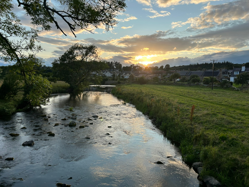 Serene river scene at sunset. A calm river flows gently through a grassy field, with scattered rocks visible in the water. In the background, a small village or cluster of houses nestles under a partly cloudy sky, the setting sun casting a warm glow. The overall mood is peaceful and tranquil.