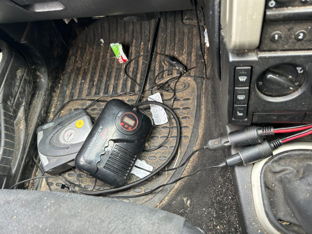 Messy interior of a car, specifically the floor area near the driver's seat. Two portable air compressors, various cords and wires, and some small debris are scattered on the floor mat.