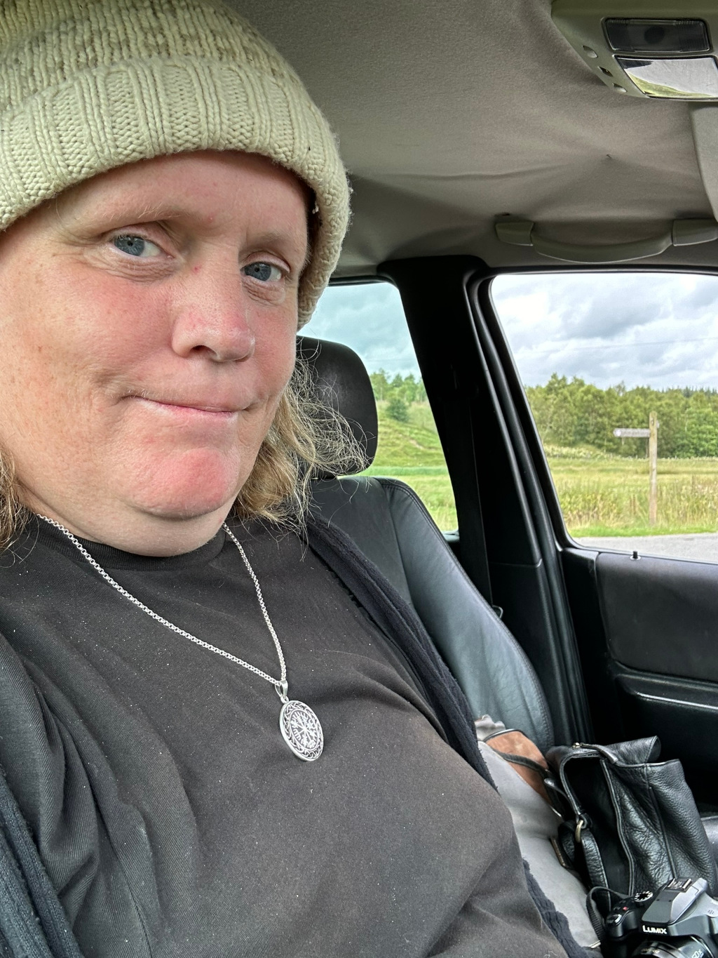 Close-up selfie of Leonie with shoulder-length blonde hair, wearing a light beige knit hat and a black shirt. She is sitting in the driver's seat of a car, looking directly at the camera with a slightly smirk. A silver necklace with a circular pendant is visible, and a black handbag and camera are partially visible in the bottom right corner. The car's window shows a rural landscape outside. The overall impression is a casual, candid self-portrait taken during a road trip or journey.