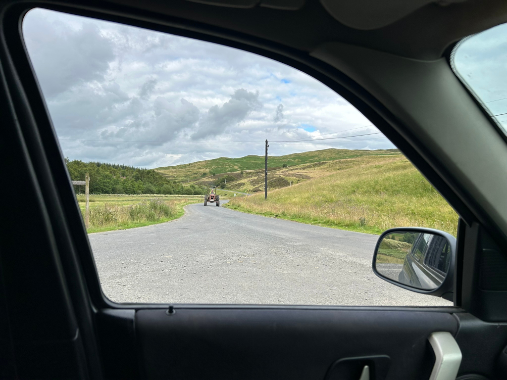 View from inside a car, looking out the window at a rural road.  A tractor is visible in the distance, travelling away from the viewer, on a paved road winding through gently rolling, grassy hills under a partly cloudy sky. The overall feeling is one of peaceful countryside scenery.