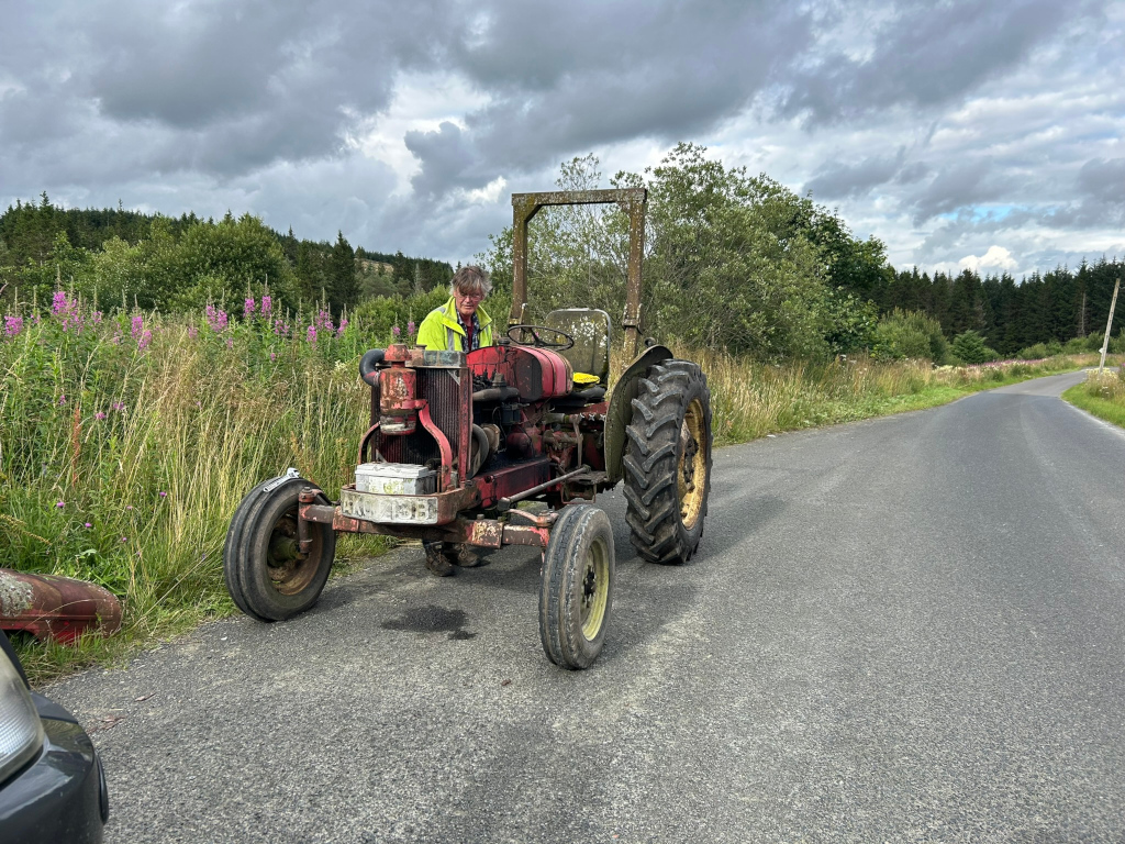 Charlie in a bright yellow safety vest sitting on an old, rusty red tractor on the side of a country road. The tractor appears well-worn and possibly in need of repair. Wildflowers and tall grass are visible along the roadside, and a dark, overcast sky hangs above. The overall scene suggests a rural setting, possibly depicting a farmer or mechanic tending to his equipment.