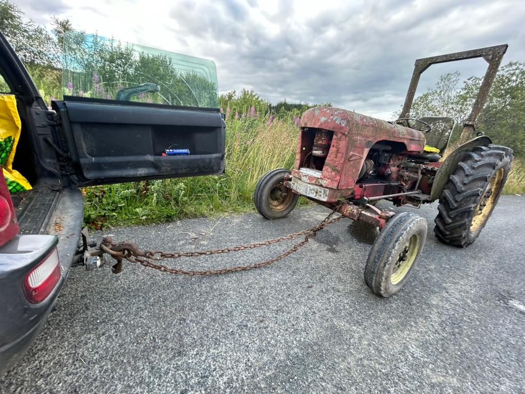 Rusty, vintage red tractor being towed by a dark-coloured SUV. The tractor appears old and weathered, while the SUV's rear hatch is open. A rusty chain connects the two vehicles. The setting appears to be a rural road with some vegetation in the background. The overall impression is one of rural life and possibly a task involving the transport of the old tractor.