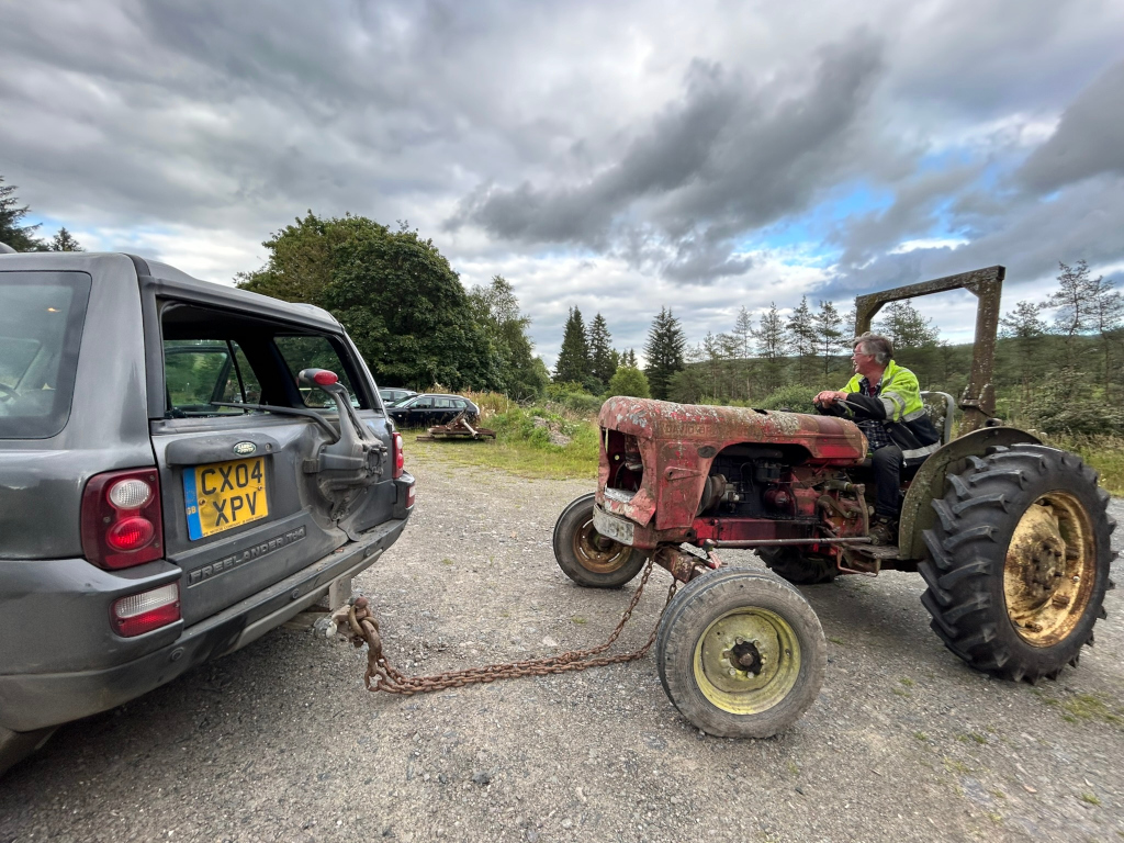 Dark grey Land Rover Freelander towing a rusty red vintage tractor along a gravel road. The tractor is driven by a man in a yellow high-visibility jacket. The background includes some trees and a cloudy sky. The scene suggests a rural setting, possibly in the UK given the license plate.
