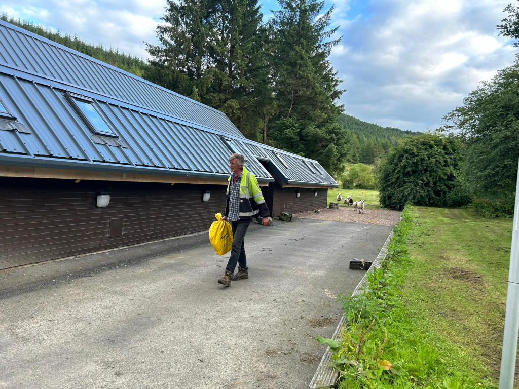 Charlie walking away from a long, dark brown building with a dark grey metal roof. The building has several skylights.  He is carrying a yellow bag. In the background, there are sheep grazing on a grassy area, and a hillside covered in trees can be seen in the distance. The overall setting appears to be rural.