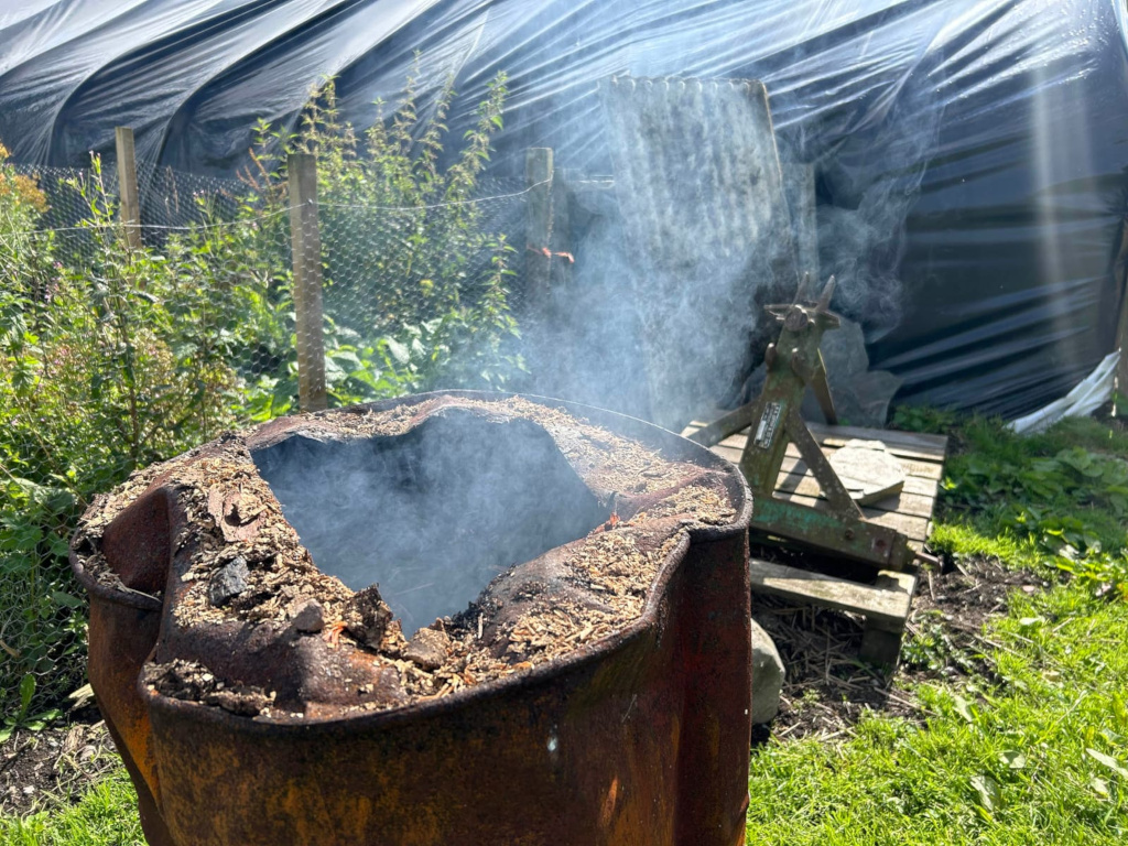 Rusty metal drum, seemingly repurposed as a makeshift smoker or fire pit. Smoke is emanating from the drum's opening, which is filled with smouldering material. The drum is situated in a garden setting, near a black plastic covering (possibly a greenhouse or garden structure) and some gardening tools or equipment. The overall scene suggests a rustic, possibly rural or agricultural, environment.