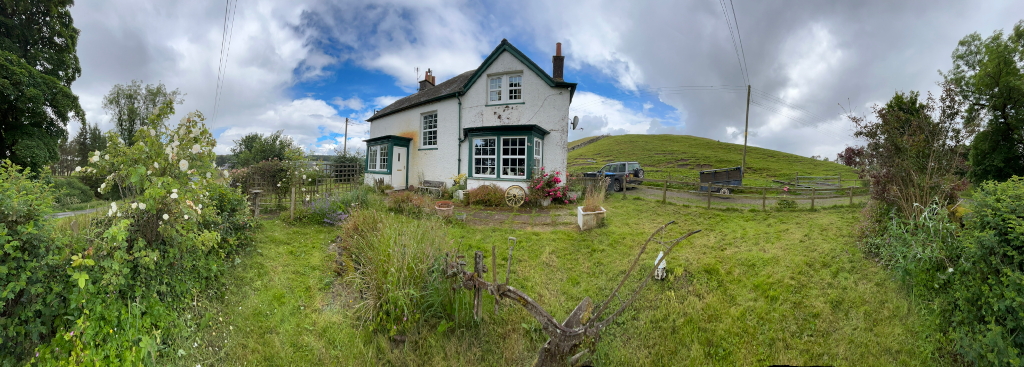 Panoramic view of a white cottage with green window frames, situated in a verdant landscape. The cottage appears somewhat aged or rustic, with an unkempt garden in front. A vehicle and trailer are parked near a fence, hinting at rural or agricultural activity. The background includes a gently sloping grassy hill under a partly cloudy sky.