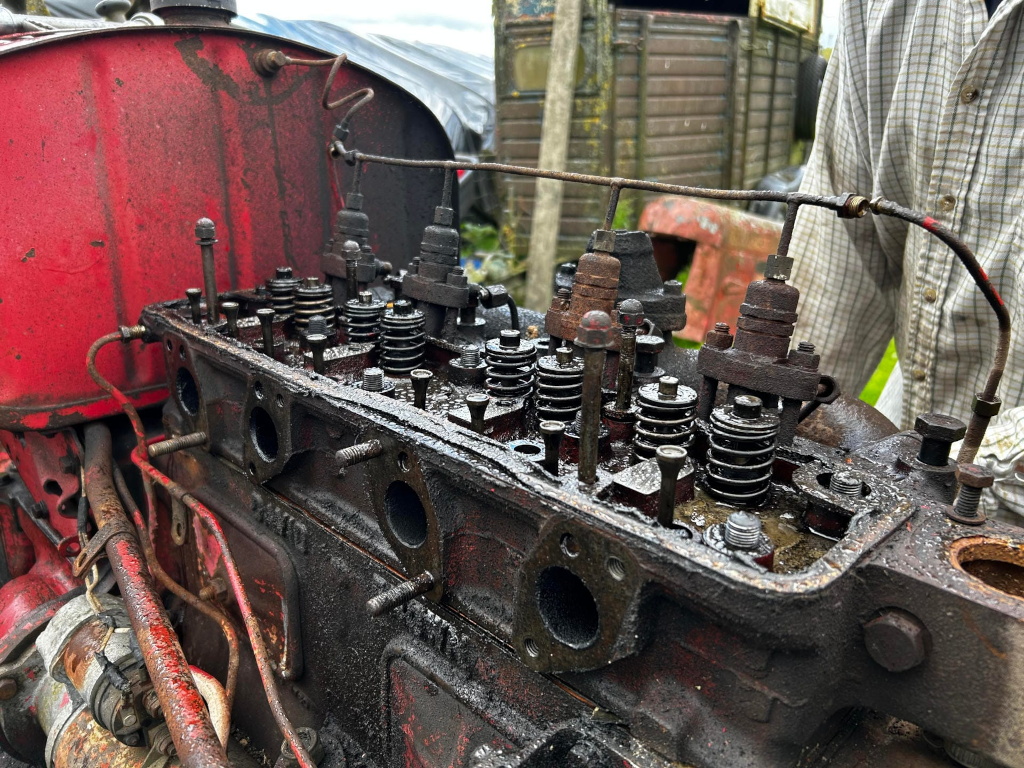 Close-up view of the disassembled cylinder head of a vintage tractor engine. The engine block is heavily soiled with oil and grime, indicating significant use and wear. The valves, valve springs, and other internal components are visible, revealing a detailed look at the engine's internal workings. The setting appears to be outdoors, in a rural or farm environment. Charlie's arm in a plaid shirt is partially visible in the background, suggesting the engine is undergoing repair or maintenance.