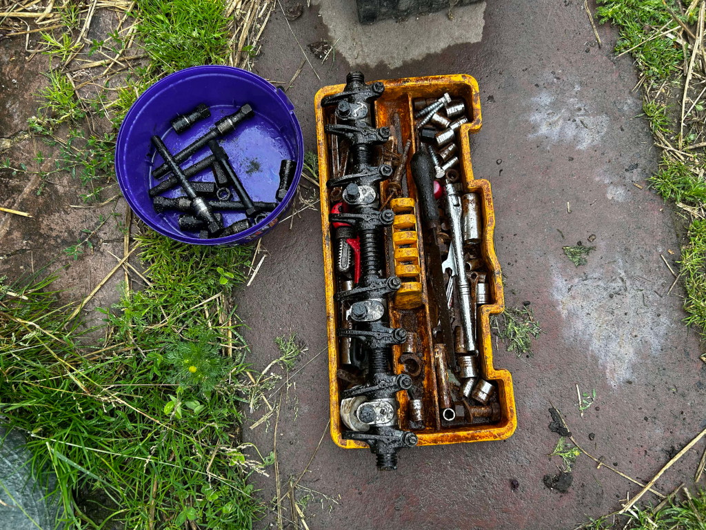 Yellow toolbox filled with various tools and parts, including a rocker arm assembly. A separate purple bucket contains a collection of dark-coloured, possibly metallic, rod-like objects. The tools and parts appear to be greasy or oily. The setting is outdoors on a patch of ground covered with grass and dirt.