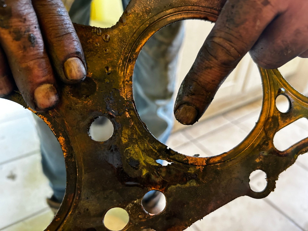 Close-up view of a damaged or worn-out engine head gasket. Charlie's dirty, grease-stained hands are holding the gasket, one finger pointing to a section of significant damage or wear. The gasket appears to be made of metal and shows signs of discoloration, oil stains, and potential blowouts or perforations. The overall impression is one of mechanical failure or the need for repair.