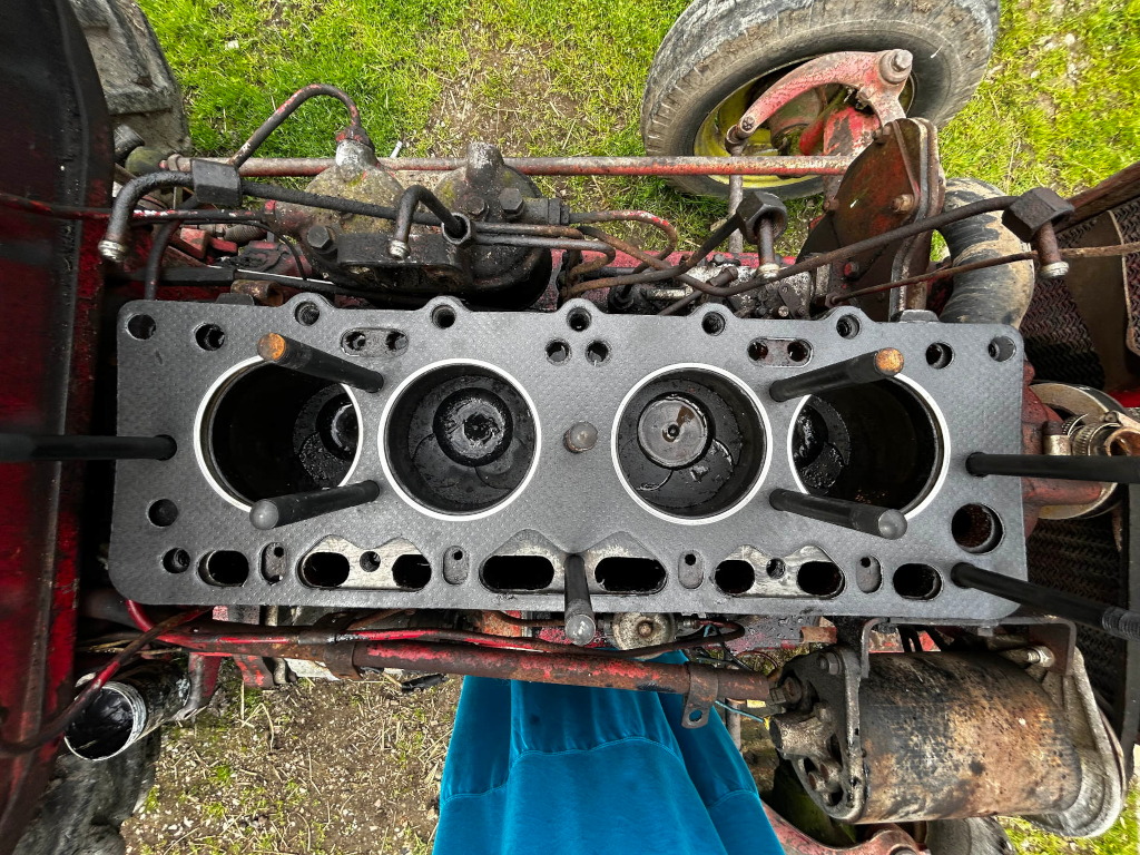 Close-up, overhead view of the engine block of a tractor. The cylinder head is removed, revealing the empty cylinder bores. The engine is visibly old and shows signs of wear and oil residue. A portion of the tractor's chassis and a wheel are also visible in the image. A piece of teal clothing is partially visible in the bottom of the frame.