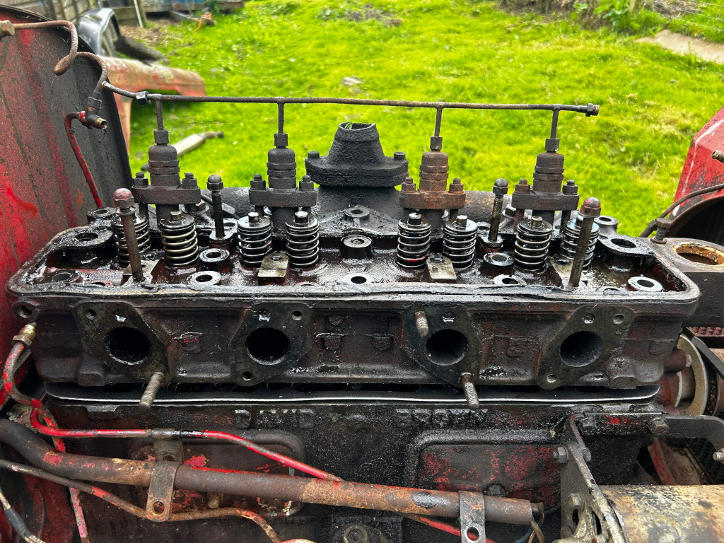 Close-up view of the disassembled cylinder head of an old, possibly vintage, internal combustion engine. The head is heavily soiled with oil and grime, indicating significant use and wear. The valves, springs, and other internal components are visible, revealing details of the engine's mechanical workings. The overall impression is one of age, use, and potential repair or restoration. The background shows a grassy area, suggesting an outdoor setting, possibly a workshop or farm.