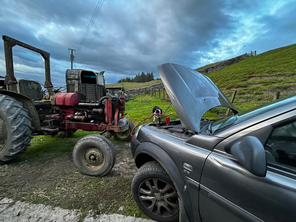 Dark grey Land Rover with its hood open, connected to an older red tractor via jumper cables. The scene is set in a rural landscape, with rolling green hills and a stone wall in the background. The overall impression is one of a roadside repair in a remote location, possibly suggesting a reliance on older, more reliable machinery in a challenging environment.