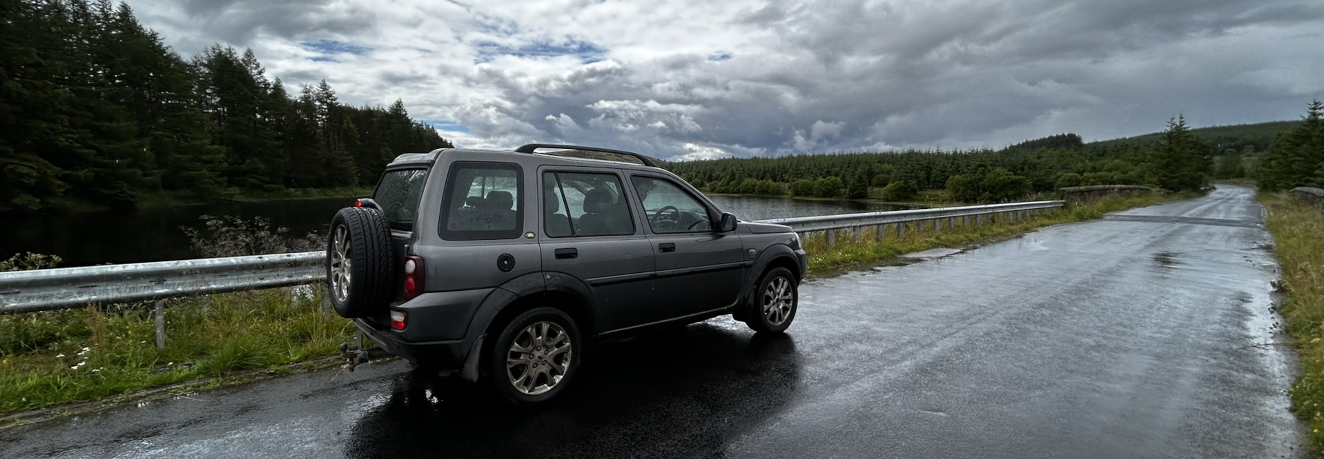 Dark grey Land Rover parked on the side of a wet road. The road is next to a body of water, and there are trees in the background. The sky is cloudy.