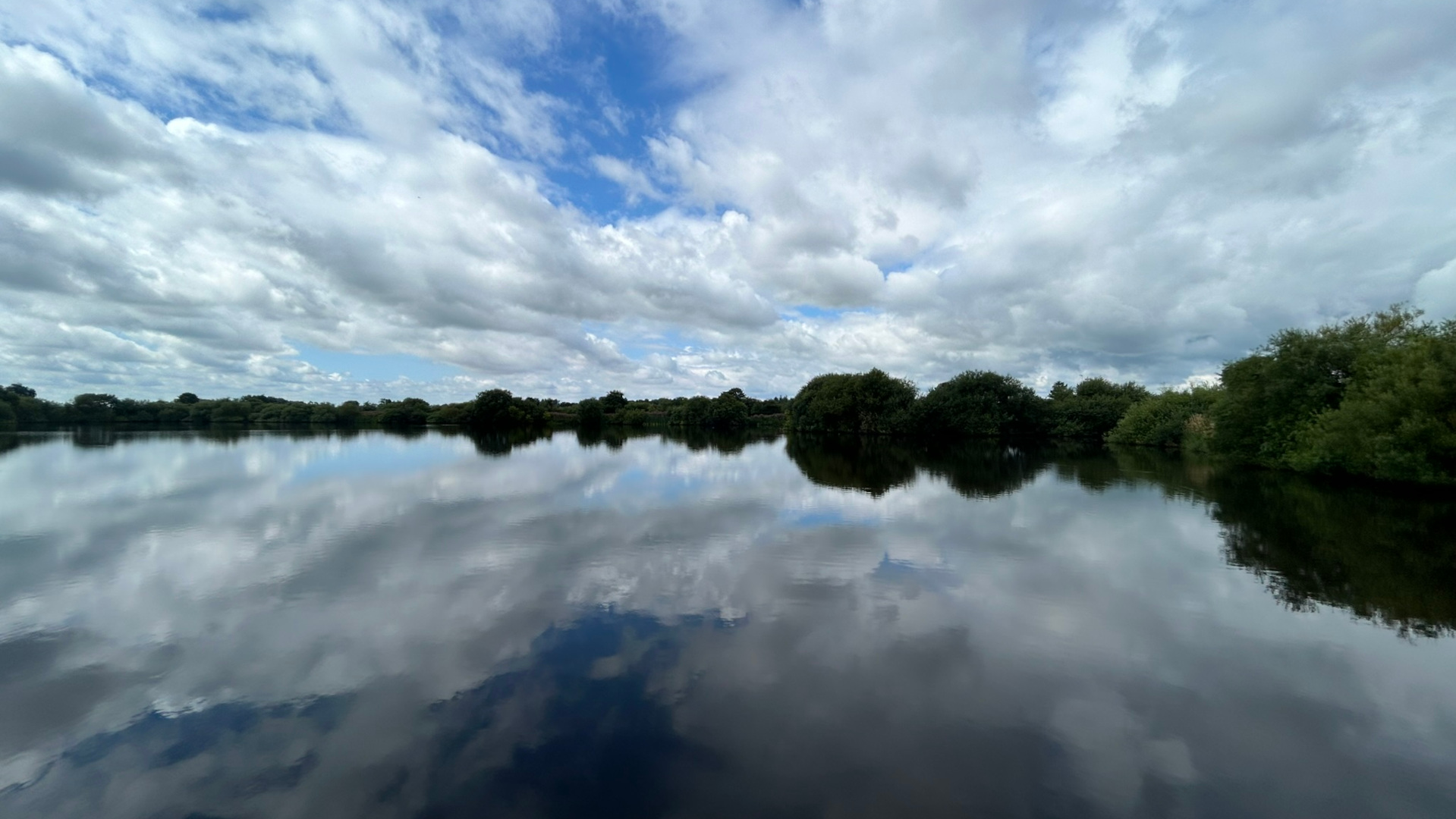Serene lake scene. The calm water perfectly reflects the cloudy sky above, creating a near-mirror image. Dark green trees and bushes line the far shore of the lake.