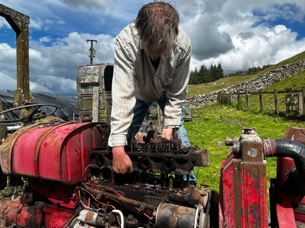 Charlie his face obscured by his long hair, is bent over an open engine compartment of a rusty red tractor. He is working on the engine, specifically the cylinder head, which is partially removed. The setting is rural, with a stone wall and grassy field visible in the background. The overall impression is one of diligent, hands-on repair work in a naturally scenic location.