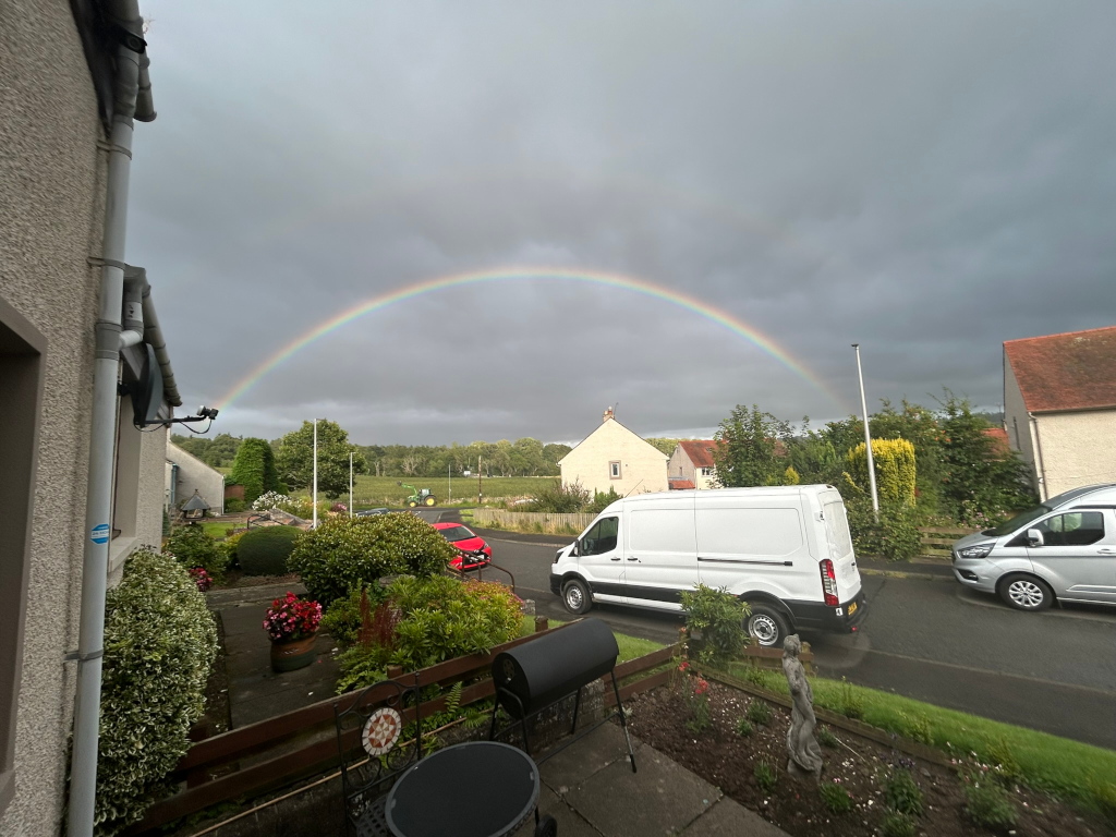 Vibrant rainbow arcing across a grey sky above a residential street. A white van is parked on the street, with a red car visible further down the road. The foreground shows a garden with bushes, flowers, and a barbecue. The overall scene is peaceful and slightly melancholic due to the overcast sky juxtaposed with the bright rainbow. The houses visible are typical of a suburban or rural setting.