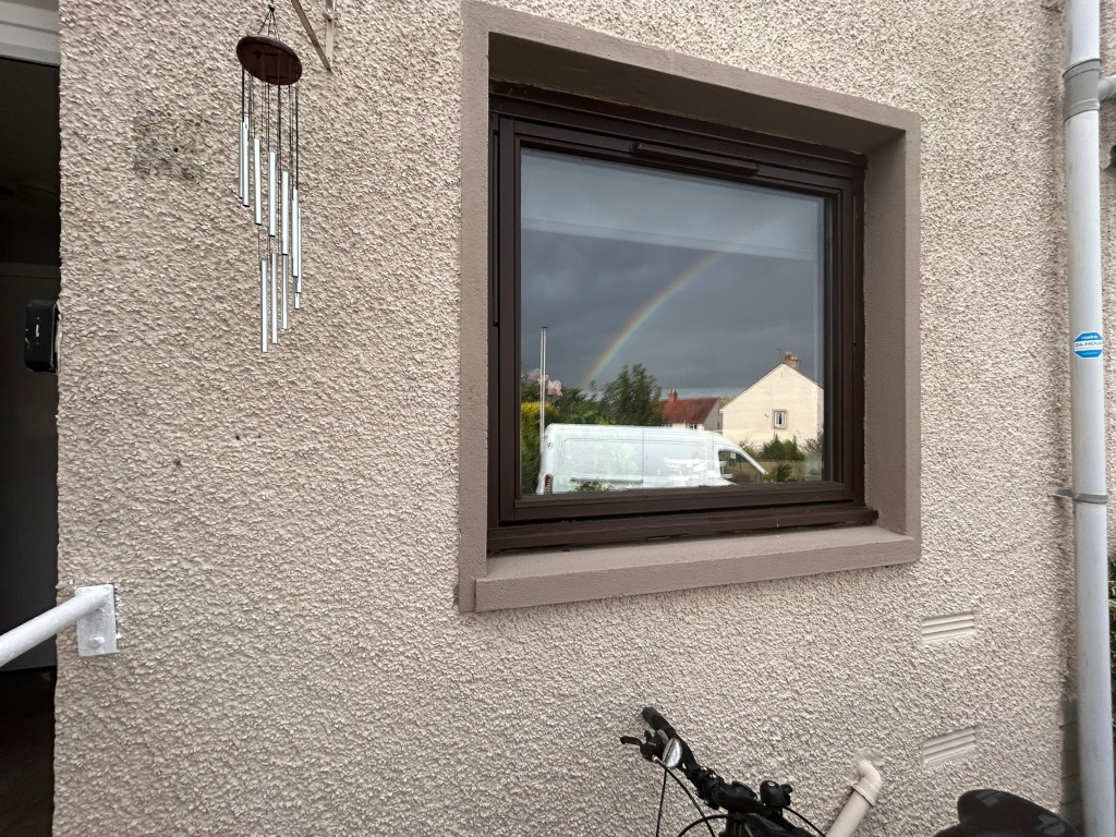 Exterior of a light beige building with a textured wall. A window reflects a rainbow and a cloudy sky, along with a white van and houses in the background. To the left of the window, a wind chime hangs on the wall. At the bottom right, part of a bicycle is visible. The overall impression is a peaceful, slightly melancholic scene, possibly after a rain shower.