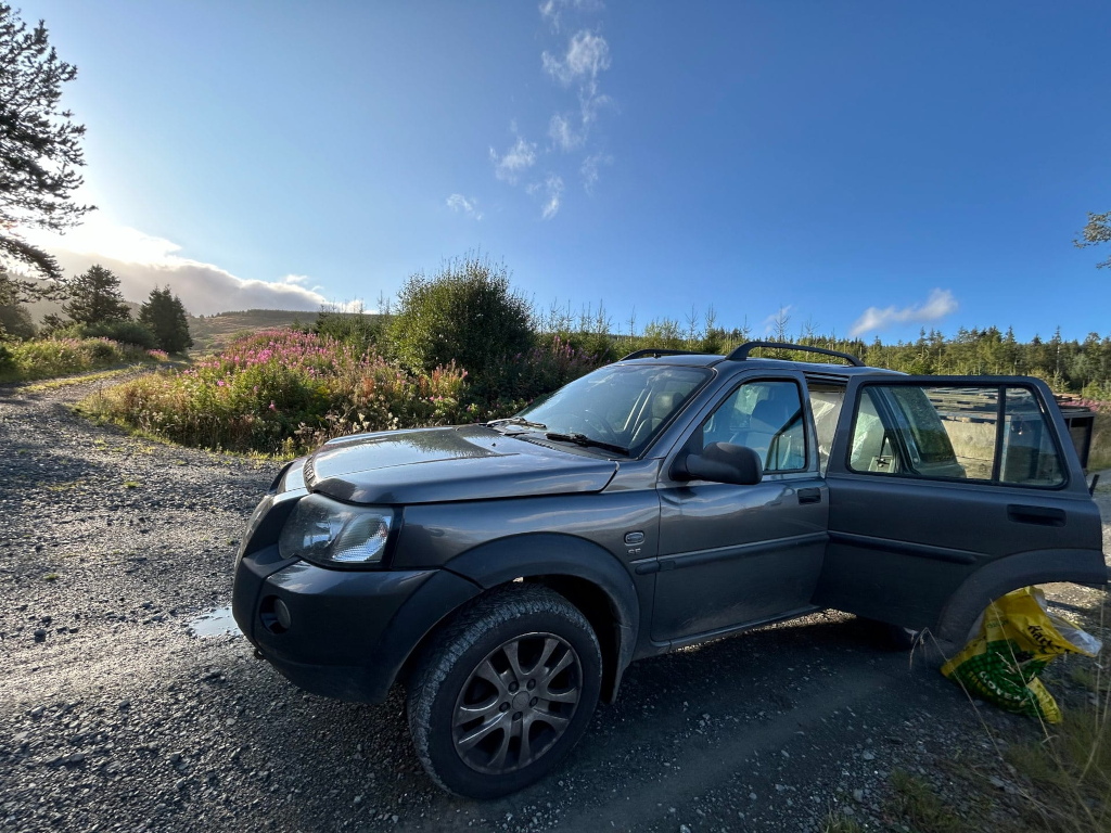 Dark gray Land Rover parked on a gravel road, with its rear passenger door open. The vehicle is situated in a rural or mountainous setting, with wildflowers and trees visible in the background. A bag of what seems to be garden fertilizer or compost is partially visible near the rear of the car, suggesting a potential gardening or landscaping activity. The sky is mostly clear and bright, indicating daytime. 