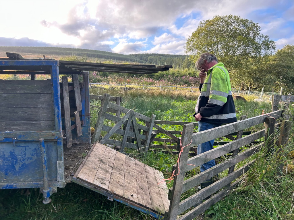 Charlie in a high-visibility jacket standing in a field, appearing pensive or troubled. He is near a dilapidated wooden livestock trailer and some broken fencing. The setting is rural, with a background of trees and hills under a partly cloudy sky. The overall mood is one of quiet contemplation and perhaps some difficulty or concern, possibly related to farm work or livestock.