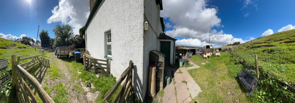 Panoramic view of a rural scene, likely in the UK. The focus is on the side of a whitewashed cottage, with a small garden and farmyard visible. Several sheep are in the background, and there's a trailer parked nearby. The overall impression is one of peaceful, rural life.