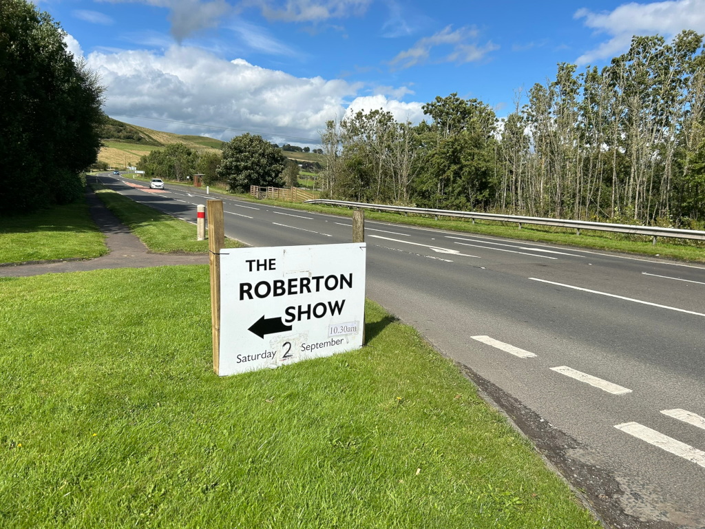 Roadside sign advertising The Roberton Show on Saturday, September 2nd, starting at 10:30 am. The sign is positioned beside a two-lane road, with green fields and trees in the background under a partly cloudy sky. The overall impression is of a rural setting leading to a local event.
