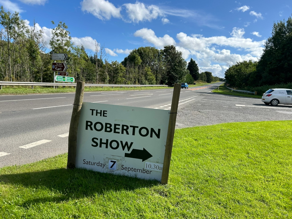 Roadside sign advertising The Roberton Show on Saturday, September 7th at 10:30 am. The sign is positioned on a grassy verge next to a two-lane road.  In the background, there are trees and a few cars are visible on the road. The overall impression is a sunny day in a rural area, with a clear indication of a local event. The image is a simple, straightforward depiction of a directional sign for a community event. There is no apparent deeper meaning or artistic expression beyond its functional purpose.