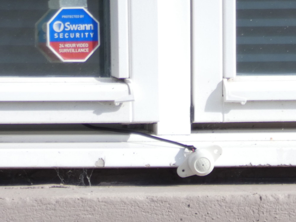 Close-up view of a window with a Swann Security sticker on it. Attached to the windowsill below is a small, white, external security sensor wired to the window frame. The overall impression is one of home security measures.